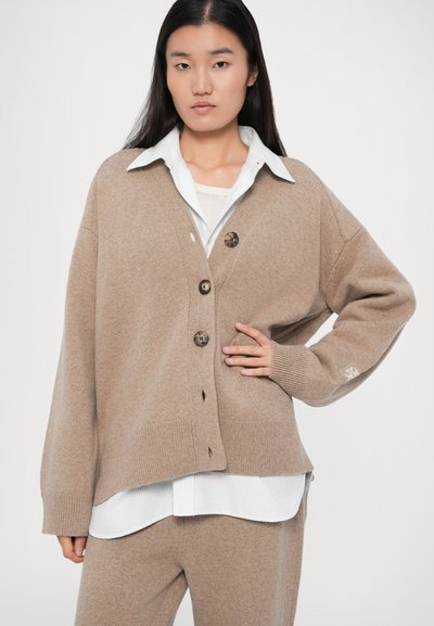 Žena v béžovém oversized kardiganu s knoflíky přes bílou košilovou halenku a odpovídající béžové kalhoty, pózující s jednou rukou na boku.