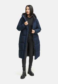 Mørkeblå lang pufferjakke med hætte, frontknaplukning og quiltet mønster. Sammen med sorte skinny jeans og sorte ankelstøvler.