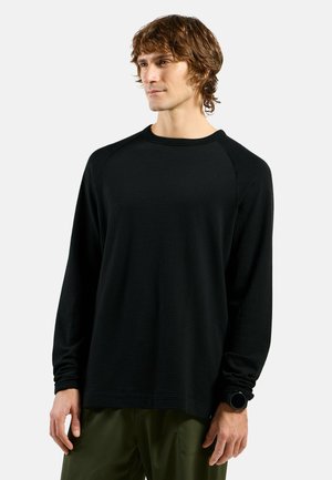 Schwarzes Langarmshirt mit strukturiertem Muster, Raglanärmeln und lockerer Passform; getragen mit grünen Hosen und einer Uhr am Handgelenk.