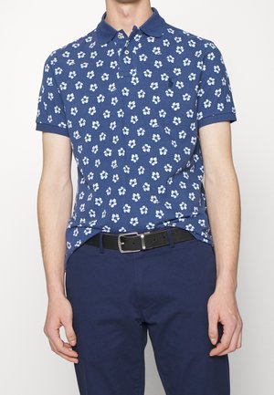 Niebieska koszulka polo z krótkim rękawem w biały kwiatowy wzór, włożona do ciemnoniebieskich spodni z czarnym paskiem i srebrną klamrą.