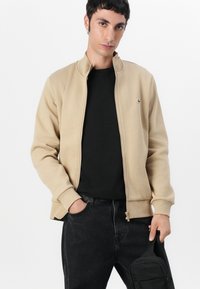 Beige zip-up jakke med ståkrage, teksturert stoff og en grønn logo på brystet. Brukt med en svart t-skjorte og mørke jeans.