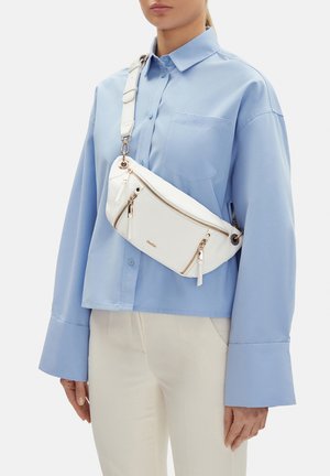 Kobieta ubrana w jasnoniebieską koszulę zapinaną na guziki i beżowe spodnie, z białą torbą na ramię z złotymi zamkami i regulowanym paskiem.