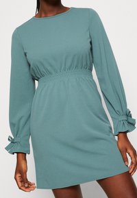 Pitkähihainen meripihkainen mekko, jossa on kerrostettu vyötärö ja solmimisnauhalla koristellut hihansuut. Pehmeä, kevyt kangas ja rento istuvuus.