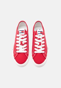 Ζευγάρι κόκκινων καμβά παπουτσιών με λευκά κορδόνια και σόλες, με το λογότυπο "Tommy Jeans" στη γλώσσα, φωτογραφημένα από πάνω σε λευκό φόντο.