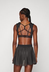 Crni sportski crop top s dizajnom s trakicama na leđima; u kombinaciji s crnom mrežastom suknjom. Sadrži prilagodljiv pojas i slojevitu tkaninu.