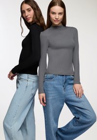 Dwie kobiety stoją plecami do siebie, ubrane w dopasowane, długie golfy w kolorze czarnym i szarym, zestawione z luźnymi niebieskimi dżinsami.