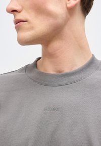 Κοντινό πλάνο στο κάτω μέρος προσώπου και στον λαιμό ενός ατόμου που φοράει γκρι φούτερ με μικρό, διακριτικό ανάγλυφο λογότυπο "HUGO" στο στήθος.