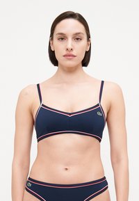Mornarska bikini zgornji del z tankimi naramnicami ter belimi in rdečimi detajli. Ima trikotno obliko in majhen zeleni logotip na levem robu.