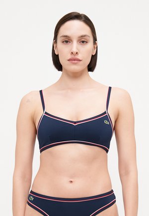 Mornarica bikini gornji dio s tankim trakama i bijelim i crvenim detaljima. Ima trokutasti oblik i mali zeleni logotip s lijeve strane.