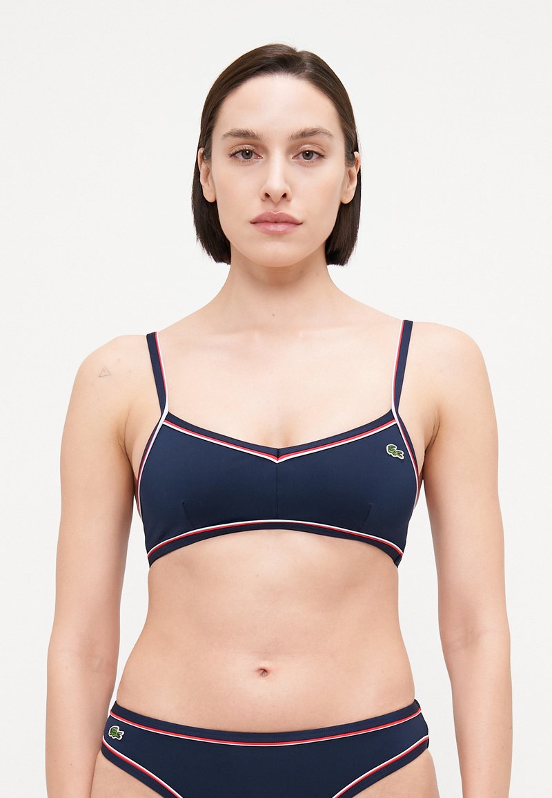 Mornarska bikini zgornji del z tankimi naramnicami ter belimi in rdečimi detajli. Ima trikotno obliko in majhen zeleni logotip na levem robu.
