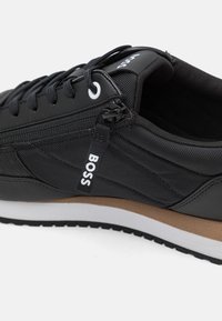 Zapatilla deportiva negra que combina materiales lisos y texturizados, con detalles en blanco y una entresuela marrón claro.