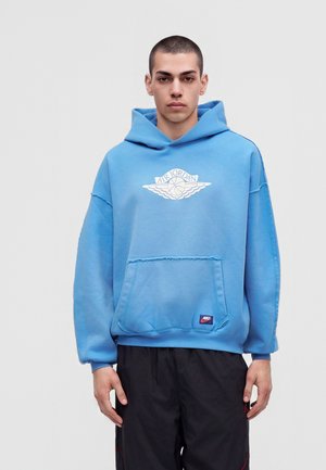 Νεαρός άνδρας με ανοιχτό μπλε φούτερ Air Jordan και μαύρο παντελόνι, στέκεται μπροστά σε απλό ανοιχτό γκρι φόντο.