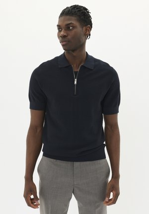 Navy short-sleeve polo shirt with a zip collar, textured fabric, and ribbed cuffs, paired with light gray pinstriped trousers.