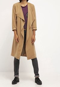 Beige langer Mantel mit drapiertem Vorderteil, aus glattem Stoff. Trägt über einem lila Oberteil, kombiniert mit grauen Jeans und schwarzen Sneakers.