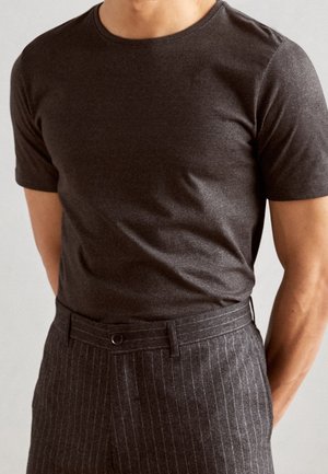 Mann trägt ein eng anliegendes dunkelbraunes kurzärmeliges T-Shirt, das in graue, fein gestreifte Hosen mit Frontknopf gesteckt ist.