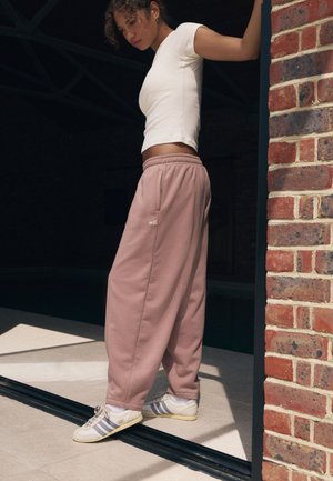 Lyserøde joggingbukser med sidelommer, elastisk talje og hvidt broderet logo, kombineret med hvide sneakers med grå striber.