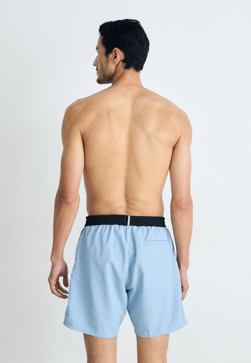 Un maillot de bain bleu clair avec une ceinture élastique noire, doté d'une poche arrière et d'un tissu lisse qui sèche rapidement.