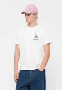 White cotton t-shirt with short sleeves and a round neckline, featuring a small colored graphic on the chest. Paired with blue denim shorts and a pink cap.