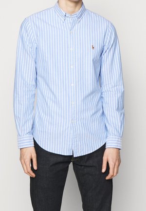 Hombre con camisa de rayas verticales en azul claro y blanco y vaqueros oscuros, de pie frente a un fondo claro y liso.