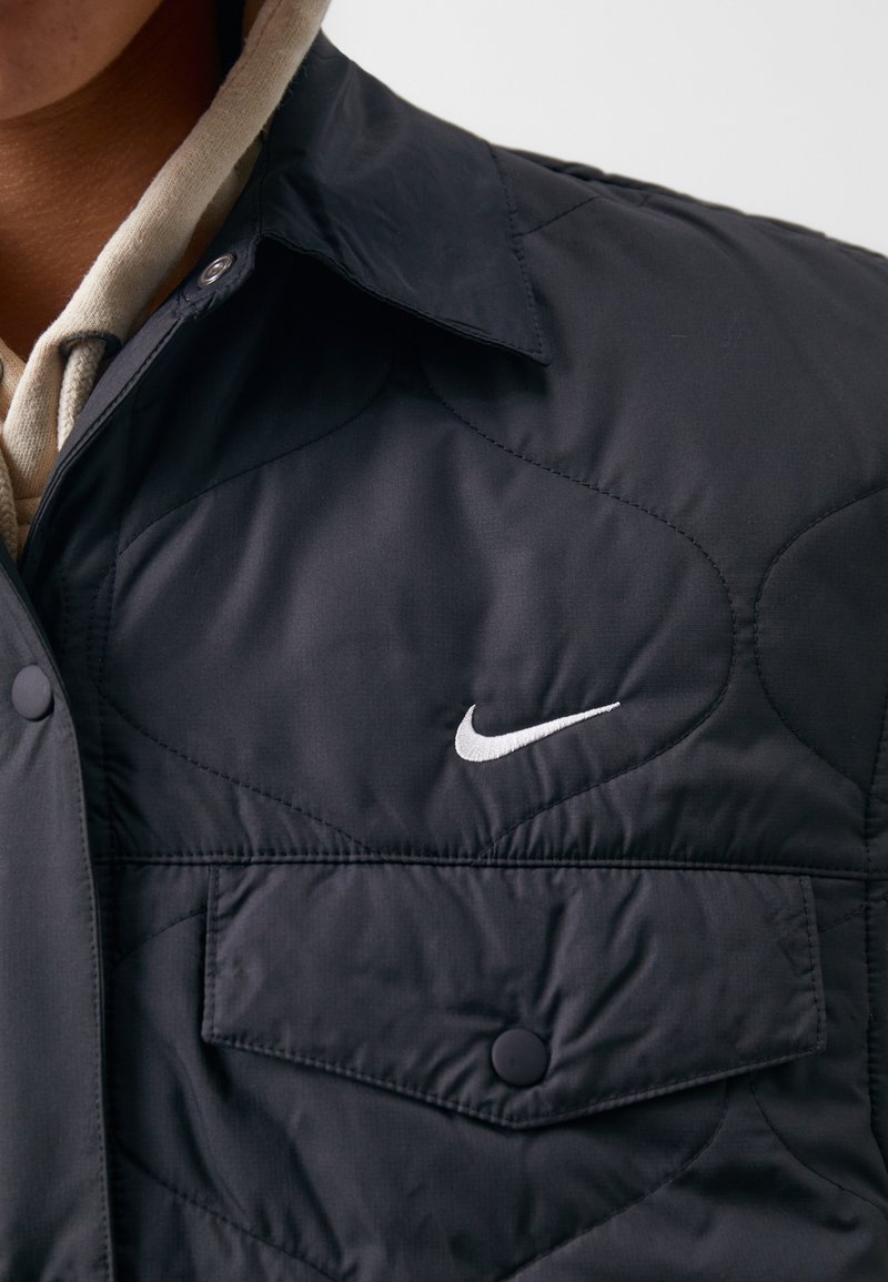 Dunkelblau gesteppte Jacke mit zwei Brusttaschen, Druckknöpfen und weißem, gesticktem Nike-Logo auf der oberen linken Seite.