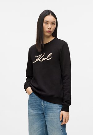 Ung kvinne med rett hår, iført en svart hettegenser med teksten "Karl" og blå jeans, stående med én hånd i lommen.