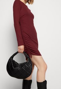 Mörkröd långärmad klänning med sidofållning, tillsammans med en svart quiltad handväska med böjd handtag och dragkedjestängning. Slät textur.