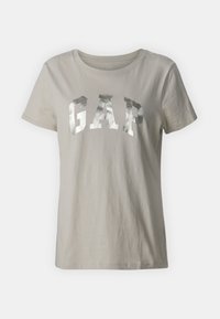 Šviesiai pilkas medvilninis marškinėlis su dideliu, metaliniu sidabriniu „GAP“ logotipu ant krūtinės. Trumpi rankoviai ir apvalus iškirpimas.