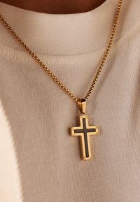 Gouden ketting met een kruispendant met zwarte accenten en decoratieve hardware, rustend op een lichtbeige gestructureerde stof.