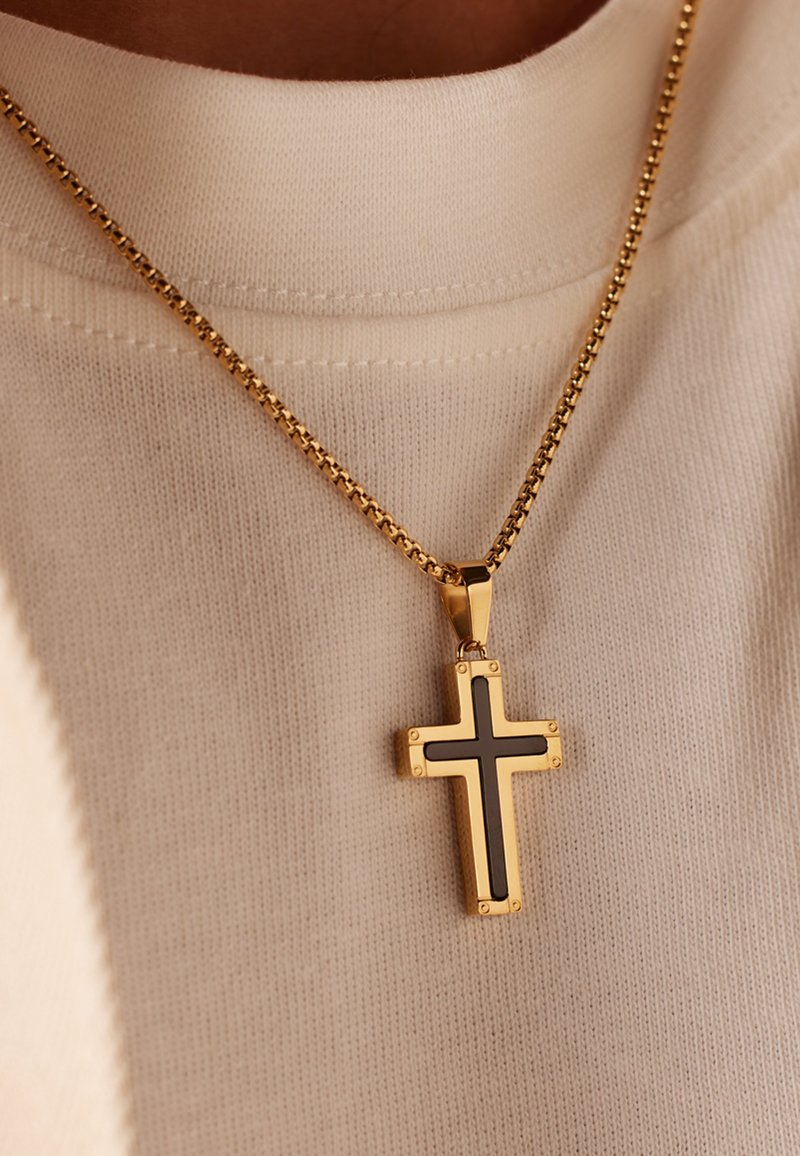 Gouden ketting met een kruispendant met zwarte accenten en decoratieve hardware, rustend op een lichtbeige gestructureerde stof.
