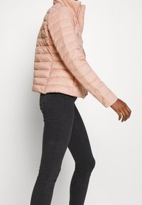 Helle rosa gepolsterte Jacke mit hohem Kragen, horizontalem Stepp-Muster und Reißverschluss vorne. Getragen mit dunklen Skinny Jeans.