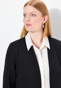 Blazer nero con spalle strutturate, abbinato a una camicia bianca con colletto. Tessuto liscio, design minimalista, lucentezza sottile e senza motivi visibili.