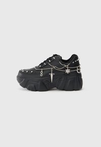 Sapatilha preta com sola chunky, adornada com correntes e charms prateados, incluindo uma adaga, sol e lua crescente. Textura suave.
