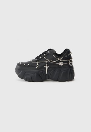 Sneaker nera con suola spessa, decorata con catene e ciondoli argentati tra cui un pugnale, un sole e una luna crescente. Texture liscia.