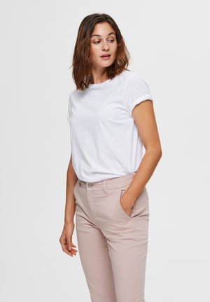 Femme aux cheveux bruns mi-longs portant une chemise blanche à manches courtes et un pantalon beige clair, regardant vers sa gauche avec une main dans la poche.