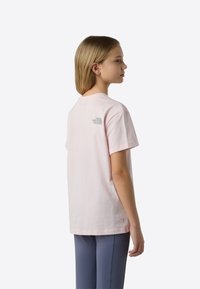 Jasnoróżowa bawełniana koszulka, krótkie rękawy, okrągły dekolt, z małym szarym logo na plecach. Noszona z niebieskimi spodniami, gładka tekstura materiału.