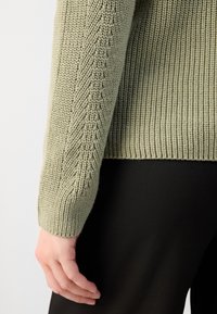 Gros plan d'une main reposant à côté du corps, portant un pull en tricot vert clair avec texture côtelée et un pantalon noir.
