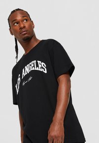 Sort bomulds T-shirt med hvid tekst "LOS ANGELES" og signaturtryk. Løs pasform med korte ærmer og rund hals.