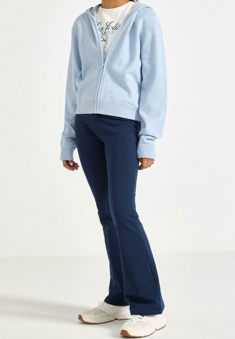Personne portant un sweat à capuche bleu clair zippé, un t-shirt blanc avec un motif graphique, un pantalon évasé bleu marine et des baskets blanches, debout devant un fond uni.