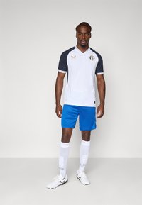 Tricou de fotbal alb și bleumarin cu logo auriu, pantaloni scurți albastre, șosete albe cu model și ghete de fotbal negre. Are un design sportiv.