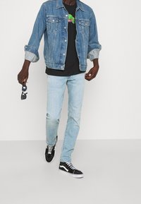 Chaqueta de mezclilla, camiseta gráfica negra, jeans azul claro y zapatillas negras con rayas blancas; sosteniendo gafas de sol en la mano izquierda.