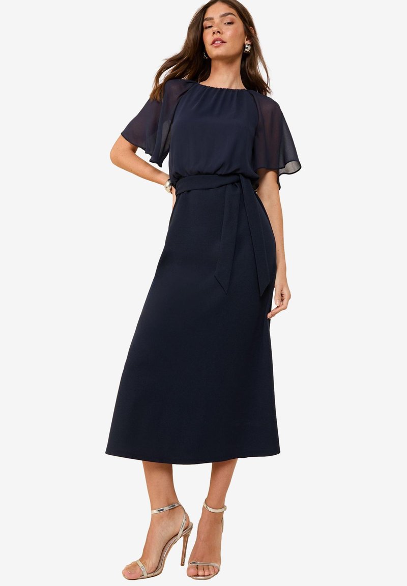 Mujer con vestido midi azul marino con mangas cortas transparentes, cinturón anudado en la cintura y sandalias plateadas de tacón alto, posando con una mano en la cadera.