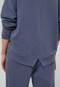 Modrá texturovaná mikina se širokými rukávy, žebrovanými manžetami a detailem s děleným lemem, doplněná o ladící spodní díl v uvolněném střihu.