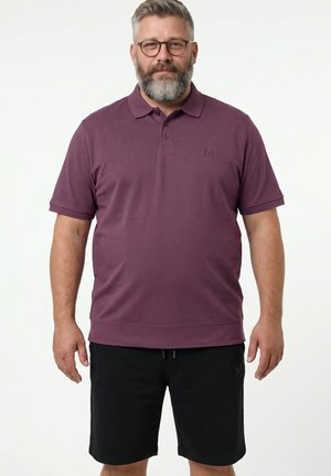 Skægget mand med briller iført en lilla polo med korte ærmer og sorte shorts, der står mod en ensfarvet hvid baggrund.