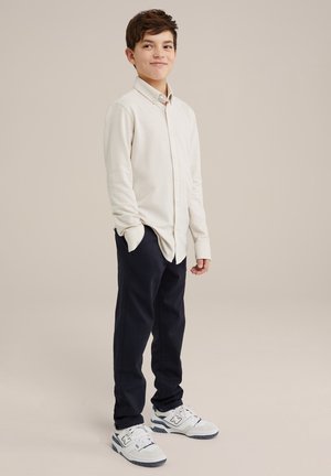 Chemise à manches longues beige clair, pantalon bleu marine et baskets blanches. Coupe décontractée, col boutonné et tissu texturé.