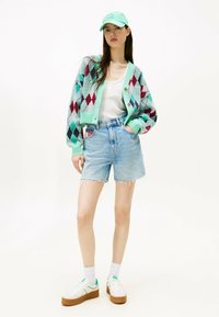 Cardigan con motivo a rombi multicolore, canotta bianca, pantaloncini di jeans e sneakers bianche con dettagli verdi. Cappellino verde chiaro.