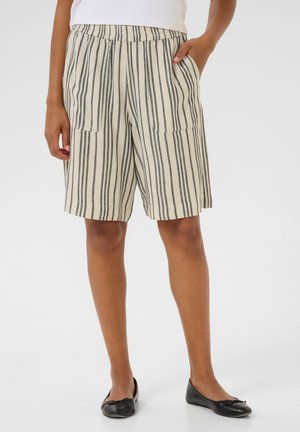 Kvinde iført beige shorts med lodrette sorte striber og forlommer, kombineret med sorte ballerinaer og en hvid top.