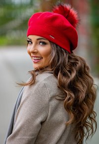 Berretto a maglia rosso con un pom-pom soffice sulla sommità, abbinato a un cappotto beige. Texture morbida e forma arrotondata. Presenta un accento del marchio metallico.