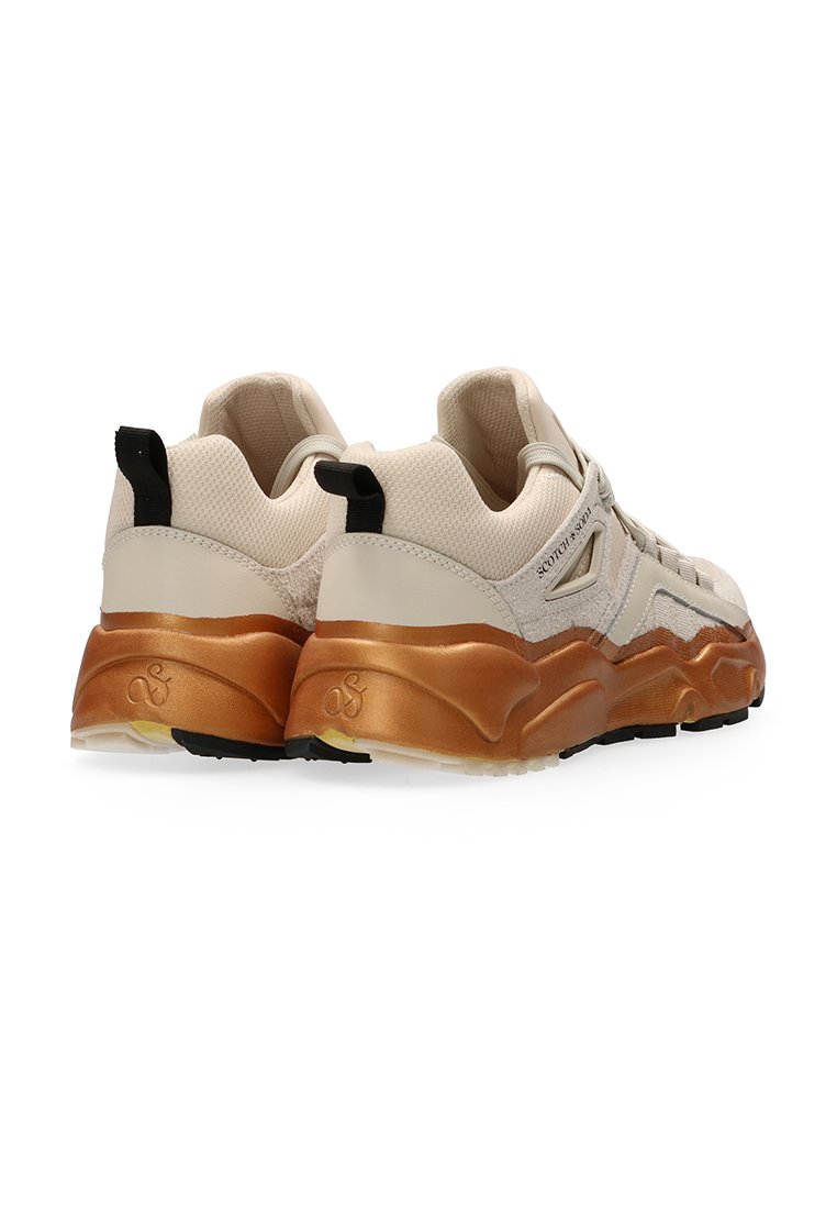 Une paire de baskets beiges et blanches avec des semelles épaisses en bronze métal, des languettes noires au talon et des tiges en maille respirante, inclinées pour montrer l'arrière et les côtés.