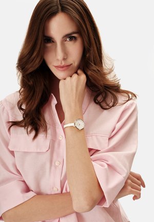 Ung kvinde med brunt hår iført en lyserød skjorte, der hviler hagen på knytnæven og viser et hvidt armbåndsur på sin venstre arm.