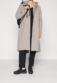 Manteau imperméable gris clair avec capuche, cordons de serrage réglables, boutons-pression, poches latérales, porté sur un haut noir et un pantalon noir, avec des chaussures de randonnée.